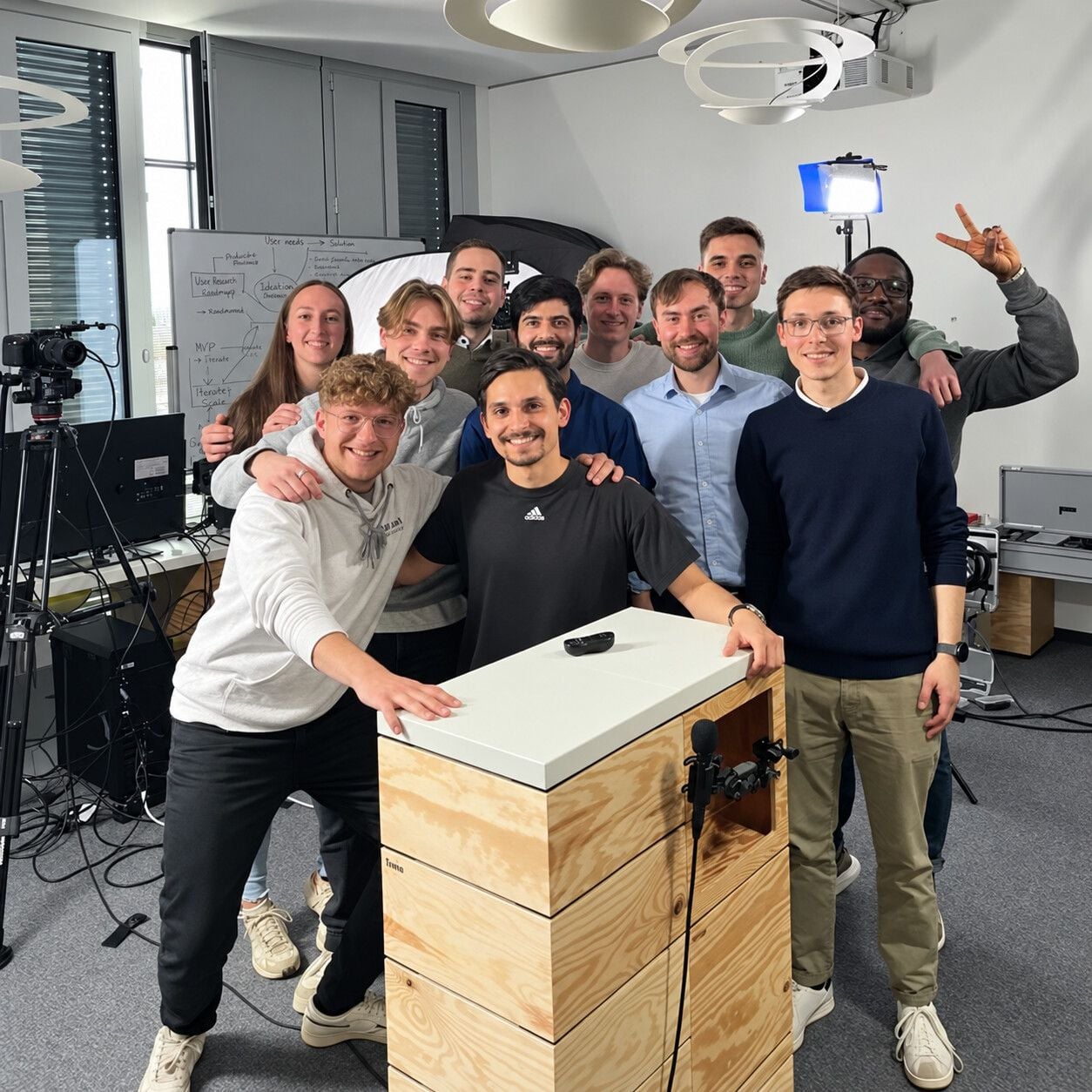 Eleven people from the Makeathon smiling around a wooden lectern with a microphone, a camera and studio light beside them