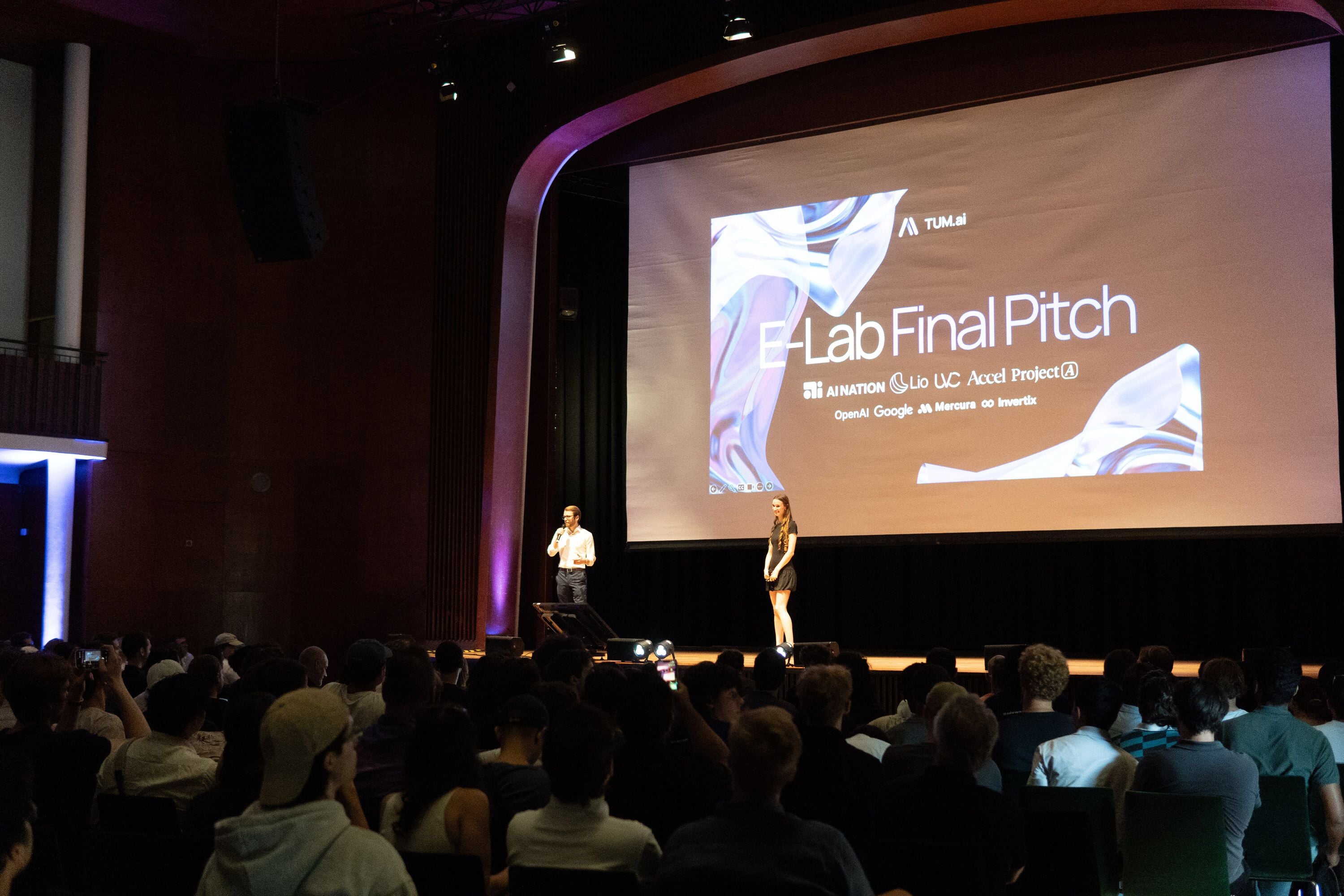 Two hosts on stage in front of a full hall at the ELAB Final Pitch, partner logos on the screen